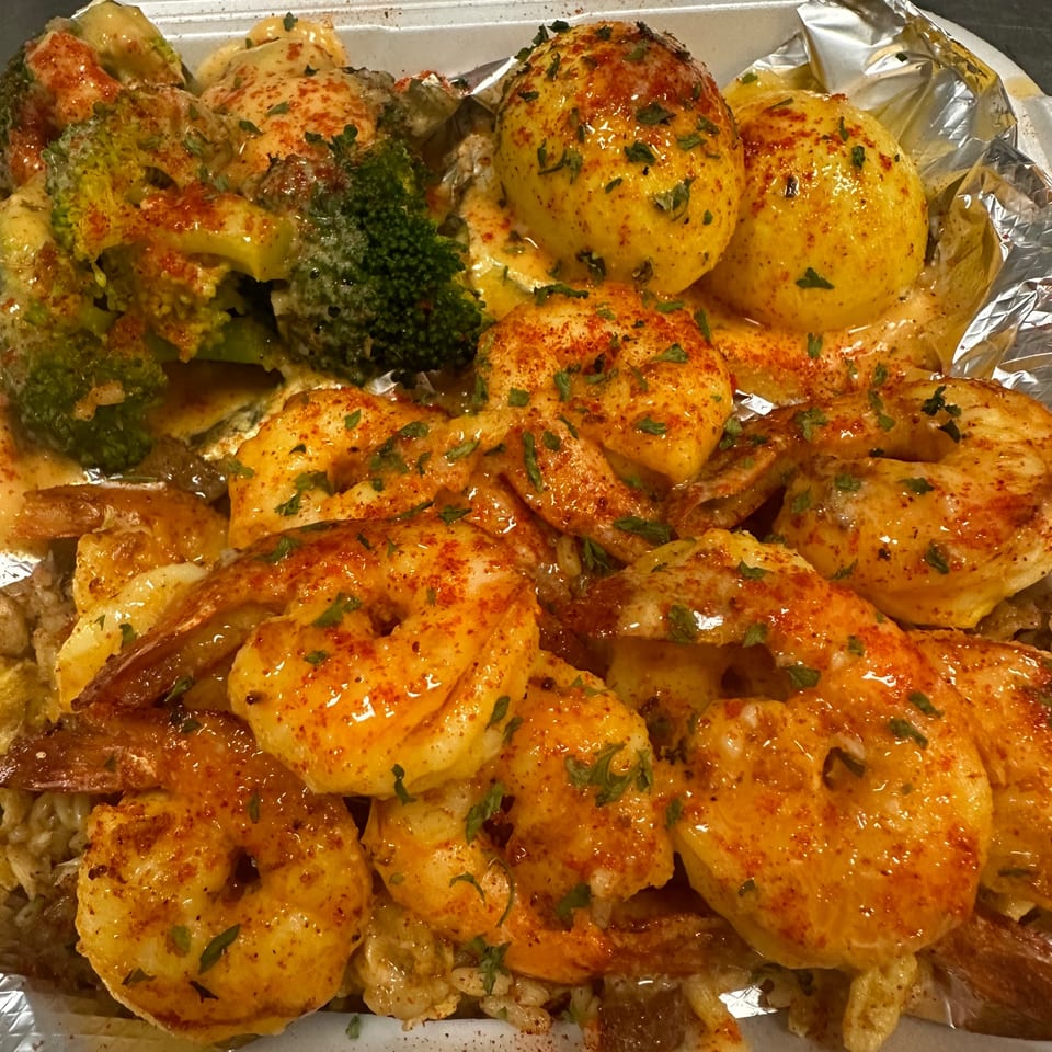 Shrimp, broccoli, and potatoes covered in sauce and seasoning, served in a foil-lined container.