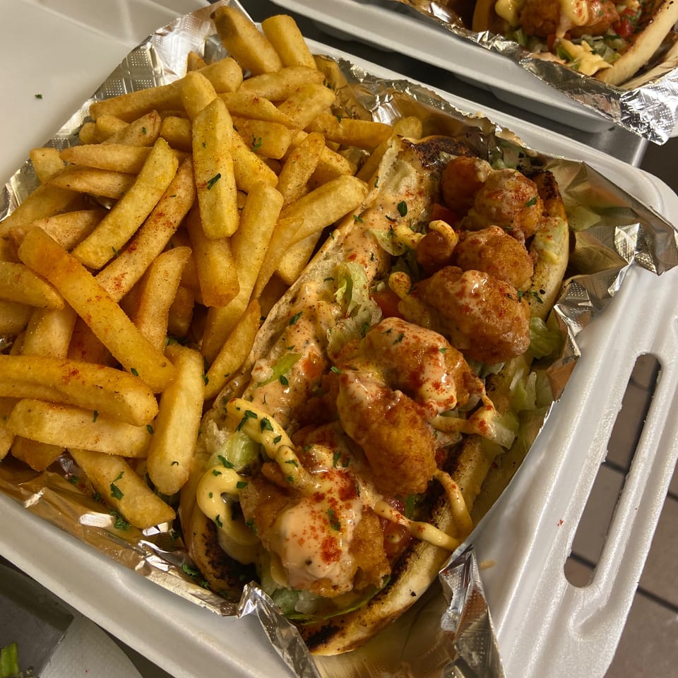 Shrimp po'boy sandwich with lettuce, sauce, and a side of seasoned french fries in a takeout container.