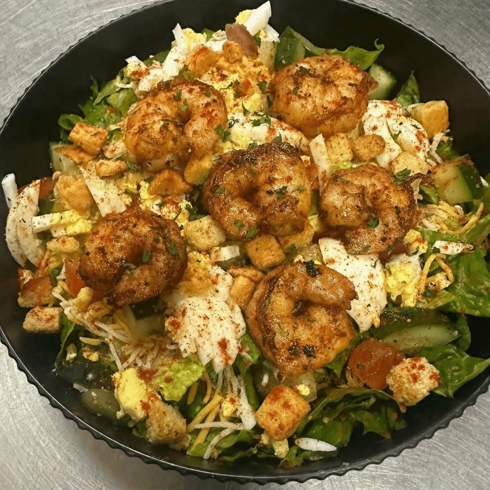 A vibrant salad with grilled shrimp, croutons, cheese, and eggs in a black bowl.