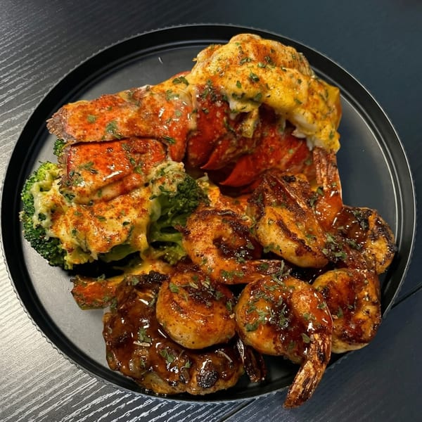 A black plate holds a lobster tail, cheesy broccoli, and grilled shrimp drizzled with sauce.