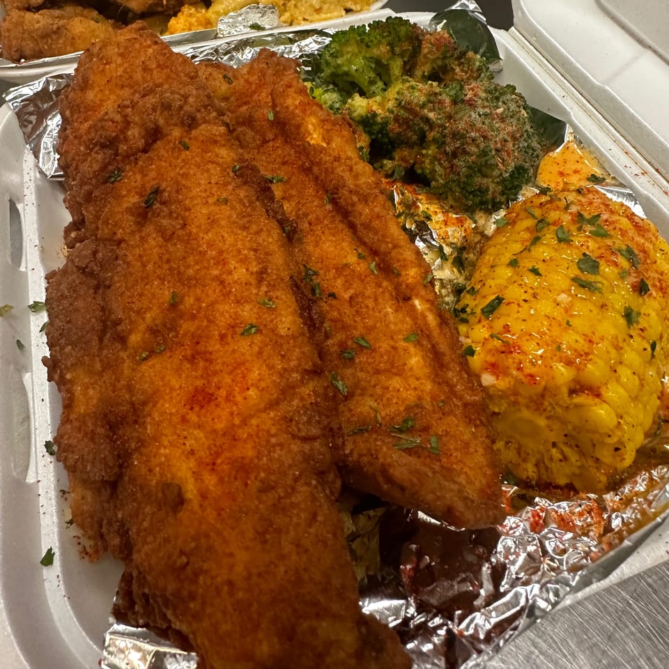 Fried fish fillets with seasoned broccoli and corn on the cob in a takeout container.
