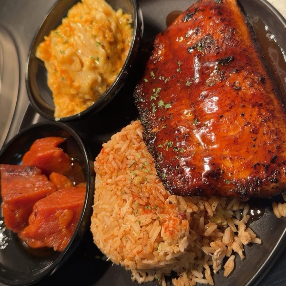 Glazed salmon with rice, macaroni and cheese, and candied yams on a black plate.